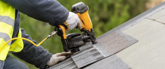 A roofer wearing a yellow vest uses a nail gun to secure shingles on a new roof.