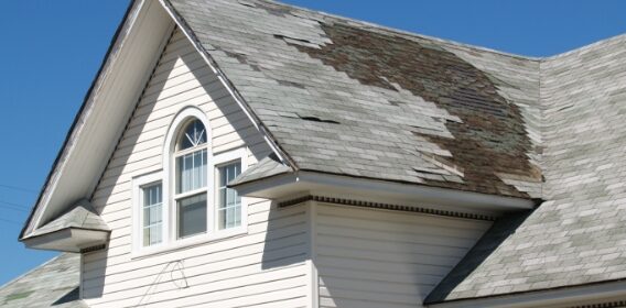 An image of a damaged grey roof on a white house, with shingles missing and baseboard exposed.