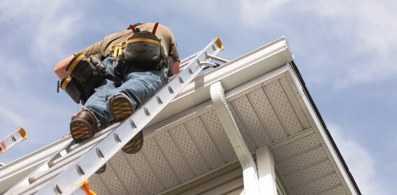 A roofer on a ladder looks at the shingles and gutter of a home.