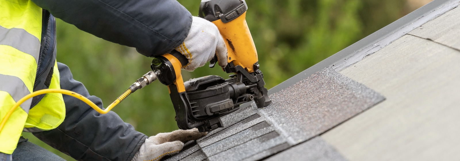 A roofer wearing a yellow vest and gloves secures shingles on a full roof replacement with a nail gun.